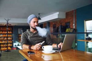 young handsome hipster man with beard sitting in cafe with a cup of coffee vaping and releases a cloud of vapor Working at laptop and having a little break